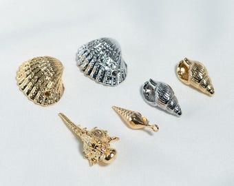 May include: An assortment of metallic seashell charms in gold and silver tones. The collection includes various shell shapes and sizes, perfect for jewelry making or craft projects. The shells are arranged on a white surface.