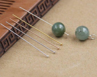 May include: A collection of four metal headpins in silver, gold, and rose gold tones, alongside two smooth, round, translucent green beads. One bead is threaded onto a silver headpin, while the other features a small silver loop at the top.