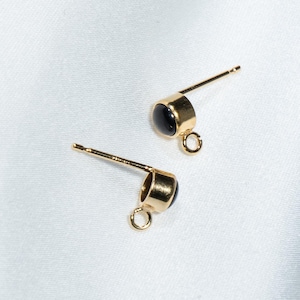 May include: Two gold-colored stud earrings with black circular stones. Each earring has a post for insertion and a small loop at the base. The earrings are set against a white, slightly textured background.