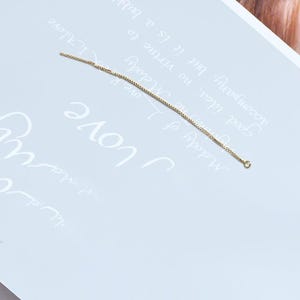 Könnte beinhalten: Ein einzelner goldfarbener Ohrring mit Kettenmuster. Der Ohrring ist dünn und länglich, mit einer kleinen Schlaufe an einem Ende und einem geraden Stift am anderen. Der Hintergrund ist eine hellblaue Oberfläche mit kursivem Text.