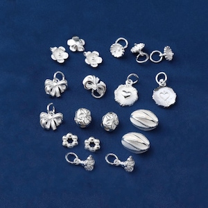 May include: A collection of small, silver-colored jewelry components on a dark blue background. The components include flower-shaped beads, bow-shaped charms, heart-shaped pendants, and oval beads. The pieces are highly detailed and reflect light.