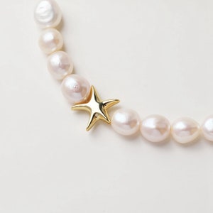 Op de afbeelding: Een parelketting met grote, crèmekleurige parels en een goudkleurige sterbedel. De parels hebben een natuurlijke, onregelmatige vorm. De sterbedel is een vijfpunts ontwerp met een gepolijste afwerking. De achtergrond is wit.