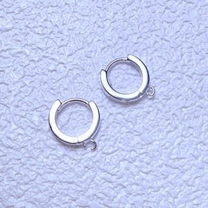 Puede incluir: Dos pendientes de aro plateados con un diseño sencillo y pulido. Cada pendiente tiene un cierre de bisagra y un pequeño lazo para sujetar abalorios. Los pendientes se muestran sobre una superficie texturizada de color blanco roto.