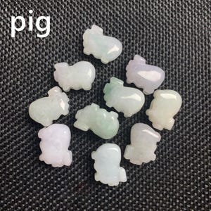 Pode incluir: Uma cole&ccedil;&atilde;o de pequenas figuras de porcos esculpidas em tons de verde, branco e lavanda. A palavra "pig" est&aacute; escrita em branco no canto superior esquerdo. As figuras est&atilde;o dispostas sobre uma superf&iacute;cie texturizada cinza escuro.