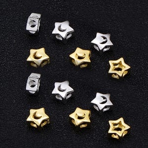 May include: A collection of star-shaped beads in silver and gold tones, some with a crescent moon cutout. The beads are small, with a hole for stringing. The background is a dark, textured surface. These beads are suitable for jewelry making.