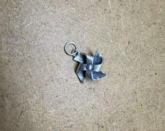 May include: A silver-colored pinwheel charm with a small loop for attaching to a necklace or bracelet. The pinwheel has four curved blades and a central rivet. The charm is set against a neutral, textured background.