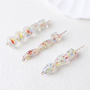 May include: Three strands of clear glass beads with colorful speckles of red, yellow, and blue. The beads are in various shapes, including round, square, and elongated, strung on thin metal wires. The beads are on a white surface.