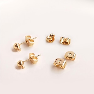 May include: A set of four gold-toned earring backs. Two of the backs are standard butterfly backs, and two are ball-shaped backs.