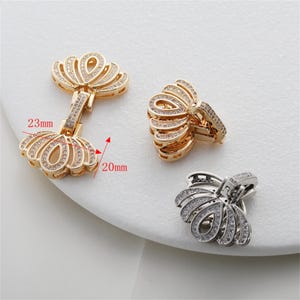 May include: Gold-toned jewelry components with a floral design, adorned with small, clear gemstones. One component measures 23mm by 20mm. The components are displayed on a white surface, showcasing their intricate details and craftsmanship.