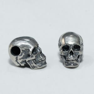 Puede incluir: Dos cuentas de metal plateado con forma de calavera y acabado pulido. Una cuenta se muestra de perfil con un orificio lateral visible, mientras que la otra se muestra de frente, revelando cuencas oculares, cavidad nasal y dientes detallados.