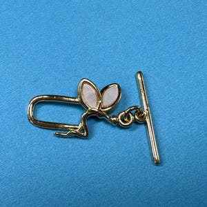Könnte beinhalten: Vergoldeter Metallverschluss mit einem dekorativen Design mit zwei weißen muschelförmigen Blättern. Der Verschluss hat eine Stange und eine Schlaufe zum Anbringen an einer Halskette oder einem Armband.