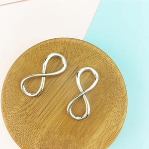 May include: Two silver infinity symbol charms on a wooden surface. The charms are smooth and shiny.