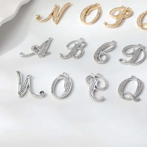 Könnte beinhalten: Nahaufnahme verschiedener Buchstabenanhänger. Einige sind goldfarben, andere silberfarben, alle mit kleinen, funkelnden Kristallen besetzt. Die Buchstaben sind in einer Schreibschrift gehalten, ideal für Schmuckherstellung oder personalisiertes Kunsthandwerk.