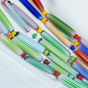 Könnte beinhalten: Eine farbenfrohe Halskette mit länglichen, gestreiften Glasperlen in verschiedenen Farben wie Grün, Blau, Rot und Gelb. Die Perlen sind mit kleinen, runden Akzentperlen aufgereiht und bilden ein lebendiges und auffälliges Accessoire.
