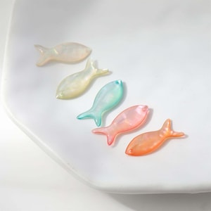 Può includere: Cinque perline iridescenti a forma di pesce nei toni del pesca, rosa, blu, giallo e crema. Le perline sono disposte diagonalmente su una superficie bianca, mostrando la loro qualità traslucida e il loro design delicato. Ideale per la bigiotteria.