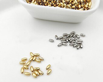 May include: A collection of small, metallic, barrel-shaped beads in gold and silver tones. A white bowl contains a large quantity of gold beads, while loose gold and silver beads are scattered on a white surface below the bowl.