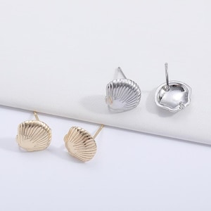 Op de afbeelding: Vier schelpvormige oorstekers. Twee zijn goudkleurig en twee zilverkleurig. De oorbellen zijn gerangschikt op een wit oppervlak, met de pinnen zichtbaar. De oorbellen hebben een getextureerd, geribbeld ontwerp.