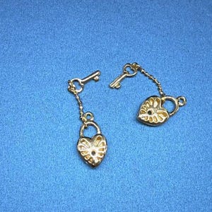 Può includere: Due portachiavi dorati con ciondoli a forma di cuore e chiavi. I ciondoli hanno un design dettagliato e sono collegati alle chiavi con una catena.