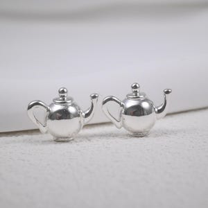 May include: A pair of shiny silver teapot-shaped stud earrings. Each earring features a small, detailed teapot design with a curved handle, spout, and a lid with a small knob. The earrings are set against a soft, neutral background.