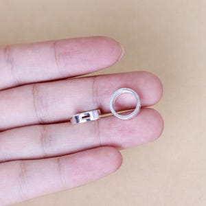 May include: Two silver jump rings, one larger than the other, are shown on a hand. The rings are smooth and have a circular shape.