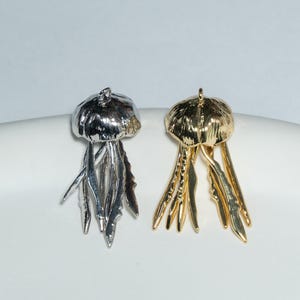 Könnte beinhalten: Zwei Quallen-Anhänger, einer silberfarben und einer goldfarben, werden gezeigt. Die Anhänger haben ein detailliertes, strukturiertes Design mit einer abgerundeten Glocke und fließenden Tentakeln. Der silberne Anhänger befindet sich links, der goldene Anhänger rechts.