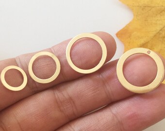 Puede incluir: Cuatro colgantes de círculo abierto de color dorado de varios tamaños. Los colgantes están dispuestos sobre un fondo claro, uno de ellos sostenido en una mano. El colgante más grande tiene un pequeño agujero. Una hoja amarilla está en el fondo.