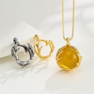 Puede incluir: Un collar de tono dorado con un colgante redondo que contiene una piedra preciosa amarilla en un marco texturizado similar a una cornamenta, junto a dos marcos de colgante vacíos en tonos plata y oro con diseños similares.