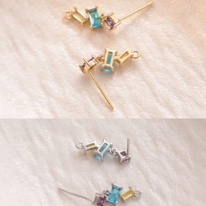 May include: Close-up of three pairs of stud earrings. Each earring features a cluster of rectangular, colorful gemstones in shades of blue, purple, and yellow. The earrings are set on gold and silver posts, displayed on a soft, white surface.