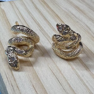 以下が含まれることがあります： クリアなラインストーンが付いたゴールドトーンのスネークイヤリングのペア。ヘビは巻き付けられており、表面にテクスチャがあります。
