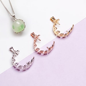 Op de afbeelding: Een collectie sieraden, waaronder een zilveren ketting met een groen-witte orb-hanger en drie halvemaanvormige oorbelcomponenten in goud, roségoud en zilver. De halvemaan oorbellen hebben een bladeren ontwerp.