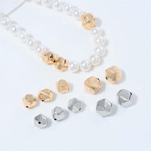 May include: A necklace with white faux pearls and gold geometric beads. Several loose gold and silver geometric beads are scattered below the necklace. The beads have a faceted design and a metallic finish. Jewelry.