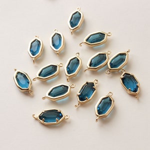 May include: A collection of gold-toned metal bezel charms with faceted, teal-blue glass inserts. Each charm has a geometric shape with a loop at each end for stringing. The charms are scattered on a light background.