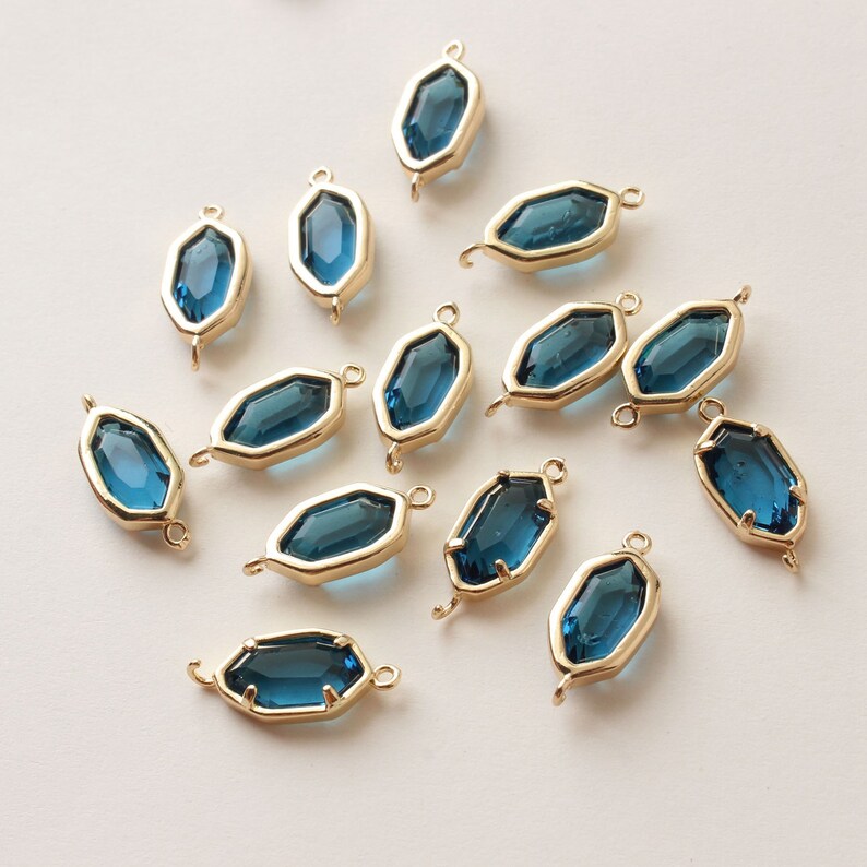 May include: A collection of gold-toned metal bezel charms with faceted, teal-blue glass inserts. Each charm has a geometric shape with a loop at each end for stringing. The charms are scattered on a light background.