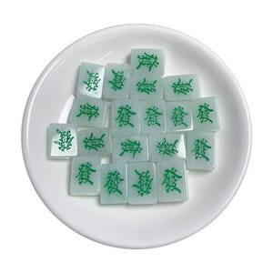May include: A white ceramic dish with a pile of green and white jade mahjong tiles. The tiles have Chinese characters on them.