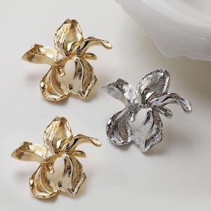 Könnte beinhalten: Drei dekorative, blütenförmige Objekte. Zwei sind goldfarben und eines ist silberfarben. Die Objekte haben ein detailliertes, strukturiertes Design, das einer Irisblüte ähnelt. Der Hintergrund ist hell und neutral.