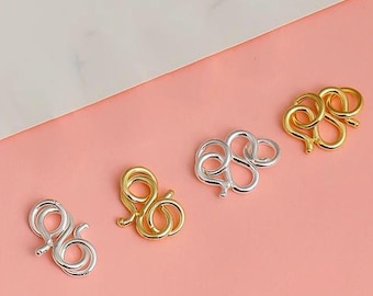 May include: Four silver and gold lobster clasps, also known as toggle clasps, on a pink background. The clasps are in the shape of a figure eight.