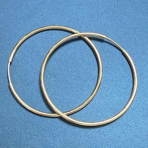 May include: Two gold-colored hoop earrings, each with a circular design. The earrings are set against a solid blue background. The hoops are thin and appear to be made of metal.