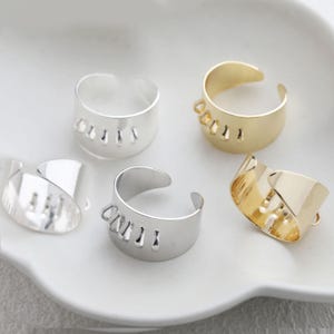 Könnte beinhalten: Fünf verstellbare Ringe in Silber- und Goldtönen werden auf einer weißen Oberfläche gezeigt. Die Ringe haben ein dickes Band mit einer Reihe vertikaler Aussparungen. Zwei Ringe sind goldfarben und drei sind silberfarben. Die Ringe sind offen für die Verstellbarkeit.