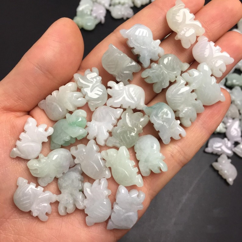 Pode incluir: Uma cole&ccedil;&atilde;o de pequenas figuras de jade esculpidas em v&aacute;rias formas de animais, incluindo tartarugas e elefantes. As figuras s&atilde;o principalmente brancas e verde-claro, com detalhes intrincados. Os objetos s&atilde;o segurados em uma m&atilde;o.