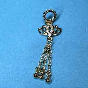 Può includere: Ciondolo dorato con un design a fiore di loto, impreziosito da cristalli trasparenti. Il gioiello presenta tre catenine pendenti, ciascuna con una piccola perla rotonda. Il gioiello è posto su uno sfondo blu.