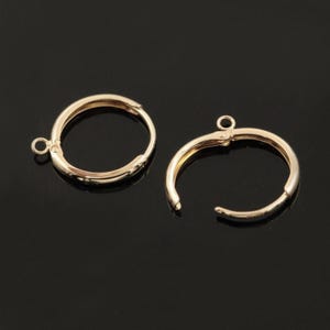 Puede incluir: Dos componentes de pendientes de aro dorados, cada uno con un diseño de bisagra y un pequeño lazo para sujetar abalorios. Los pendientes se muestran sobre un fondo negro.