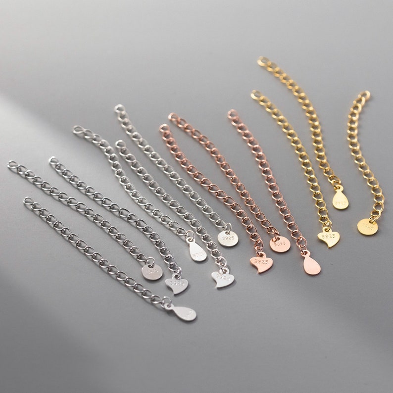 May include: A set of eight silver, gold, and rose gold chain extenders with different charms. The charms include a teardrop and a heart shape. Each charm has the number "925" engraved on it.