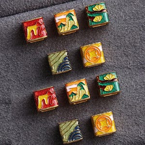 May include: A collection of small, rectangular, decorative beads with gold-colored edges. Each bead features a unique design, including landscapes, abstract patterns, and circular motifs, in vibrant colors like red, green, yellow, and blue.