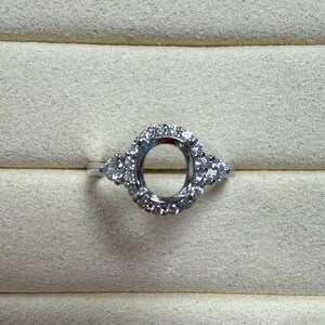 May include: A silver ring with a round, empty setting surrounded by small diamonds. The ring has a delicate design with small diamonds on the sides.