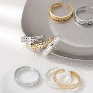 May include: A collection of open-ended rings in gold and silver hues. The rings are characterised by a series of small, evenly spaced holes along their bands. Some rings have a double band design. The jewellery is displayed on a silver-coloured, circular tray.