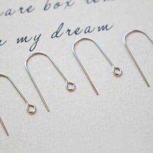 May include: Five silver earring hooks, also known as ear wires, are arranged in a row on a white surface. The hooks are all the same size and shape, with a small loop at the top and a straight wire extending down.
