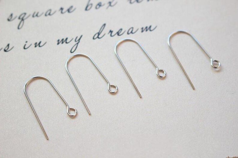 May include: Five silver earring hooks, also known as ear wires, are arranged in a row on a white surface. The hooks are all the same size and shape, with a small loop at the top and a straight wire extending down.