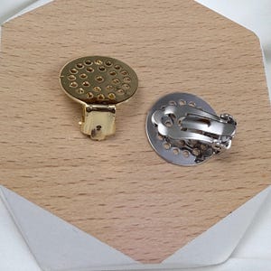 May include: Two clip-on earrings. One is gold-toned with a circular, perforated design and a hinged clip. The other is silver-toned with a similar perforated design and a clip-on mechanism. Both are displayed on a wooden surface.