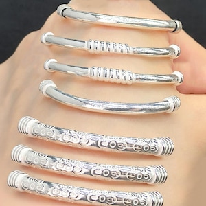 May include: A collection of silver-colored bangles. Some bangles have a smooth, curved design, while others feature a coiled or patterned central section. The bangles are displayed against a neutral background.