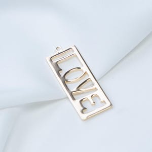 Op de afbeelding: Een goudkleurige hanger in een rechthoekige vorm, met het woord "LOVE" uitgesneden. De hanger heeft een kleine lus aan de bovenkant om aan een ketting of koord te bevestigen. De achtergrond is een witte, gevouwen stof.